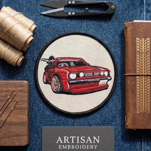 Puede incluir: Un parche bordado circular que presenta un coche deportivo rojo sobre un fondo crema con un borde negro. El parche se muestra junto a unas tijeras de hilo negras, carretes de madera con hilo beige, un estuche de madera y un diario de cuero marrón.