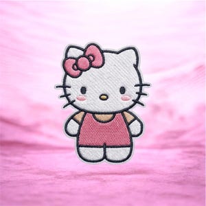 Peut inclure: Écusson brodé représentant un chat blanc avec un nœud rose, vêtu d'une robe rose. Le chat a des moustaches noires, de petits yeux noirs et des joues roses. L'écusson a une bordure blanche et est destiné aux vêtements ou accessoires.