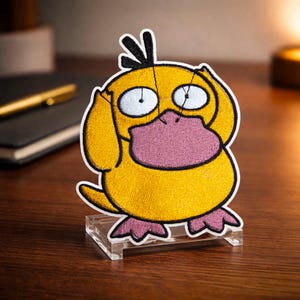 May include: Embroidered yellow Psyduck patch with a white outline, pink beak and feet, and black hair. The patch is displayed on a clear acrylic stand, set against a dark wooden surface.