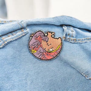May include: A circular embroidered patch depicting a stylized wave in purple, red, and yellow. The patch is attached to a light blue denim jacket. The design includes a boat and is inspired by Japanese art.