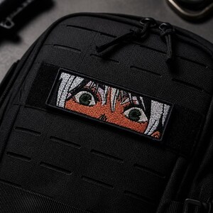 Peut inclure: Une pochette tactique noire avec un écusson rectangulaire brodé. L'écusson présente un gros plan d'yeux de style anime avec des cheveux blancs, des iris verts et une peau bronzée, sur un fond noir avec des sangles horizontales.