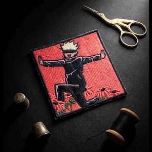 Op de afbeelding: Geborduurde patch met een personage in zwarte kleding met een blinddoek, tegen een rode achtergrond met bloemen. Gouden schaar, vingerhoeden en een spoel zwart garen zijn rond de patch op een zwart oppervlak geplaatst.