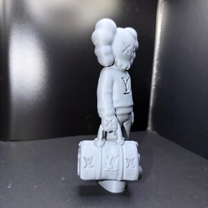 Puede incluir: Una figura gris de un personaje con los ojos tachados y una cabeza en forma de nube, que lleva un su&eacute;ter con un logotipo "Y" y sostiene una bolsa de lona gris con logotipos "LV". La figura est&aacute; de pie sobre una superficie oscura.