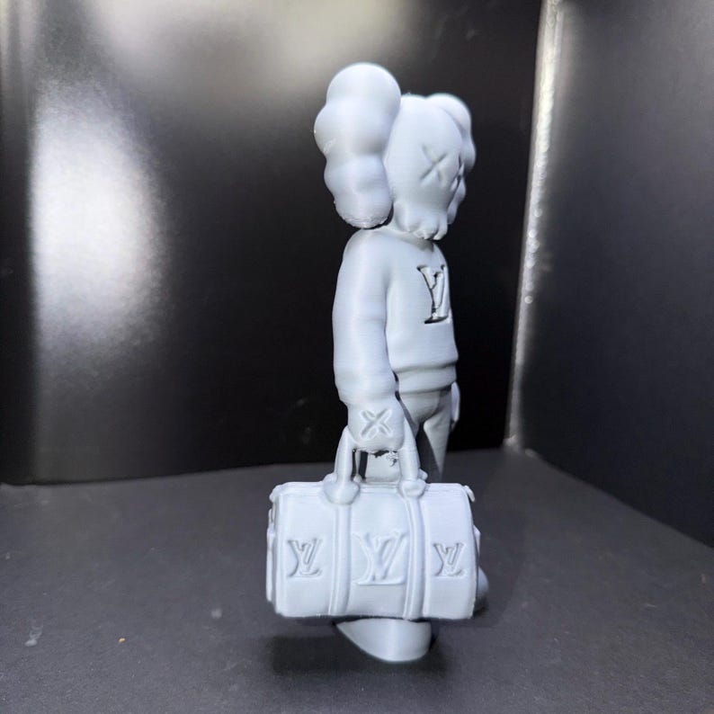 Puede incluir: Una figura gris de un personaje con los ojos tachados y una cabeza en forma de nube, que lleva un su&eacute;ter con un logotipo "Y" y sostiene una bolsa de lona gris con logotipos "LV". La figura est&aacute; de pie sobre una superficie oscura.