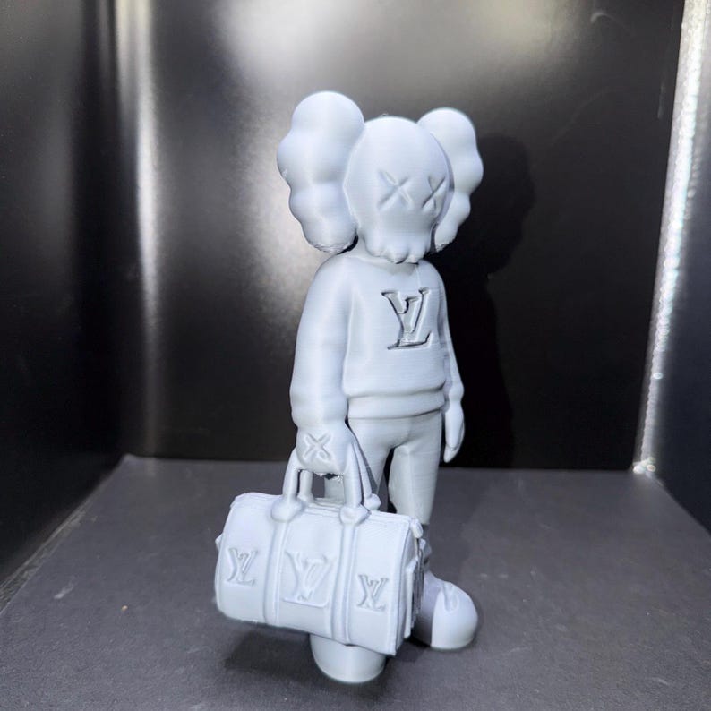 Puede incluir: Una figura gris claro de un personaje con los ojos tachados y orejas grandes, que lleva un su&eacute;ter con un logotipo y sostiene una bolsa de lona a juego. La bolsa y el su&eacute;ter tienen el mismo logotipo. La figura est&aacute; de pie.