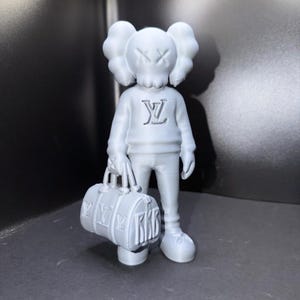Puede incluir: Figura gris claro de un personaje de dibujos animados con ojos tachados y orejas grandes, que lleva un su&eacute;ter con un logotipo y sostiene una bolsa de lona. La bolsa y el su&eacute;ter presentan el mismo logotipo. La figura est&aacute; sobre una superficie gris.