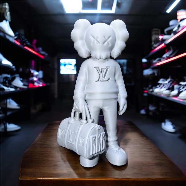 Puede incluir: Una figura blanca de un personaje de dibujos animados con los ojos tachados, que lleva un su&eacute;ter con un logotipo y sostiene una bolsa de lona. La bolsa tiene las letras "RKB" y un logotipo. La figura est&aacute; sobre una superficie de madera.