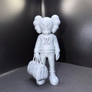 Puede incluir: Una figura gris de un personaje de dibujos animados con una forma de cabeza distintiva y ojos tachados. La figura lleva un su&eacute;ter con un logotipo y sostiene una bolsa de lona. La bolsa tiene las letras "RKB".