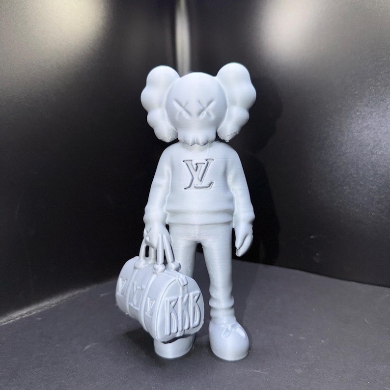 Puede incluir: Una figura gris de un personaje de dibujos animados con una forma de cabeza distintiva y ojos tachados. La figura lleva un su&eacute;ter con un logotipo y sostiene una bolsa de lona. La bolsa tiene las letras "RKB".