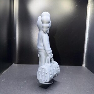 Puede incluir: Figura gris claro de un personaje de dibujos animados sosteniendo una bolsa de lona. La bolsa tiene las letras "KKK" y "LV". El personaje lleva un su&eacute;ter y tiene una forma de cabeza distintiva. Objeto decorativo.