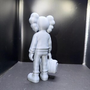 Puede incluir: Figura gris claro de un personaje de dibujos animados con orejas grandes y redondeadas y una cabeza redondeada. La figura lleva un su&eacute;ter, pantalones y zapatos, y sostiene una peque&ntilde;a maleta. La figura est&aacute; de pie frente a un fondo oscuro.