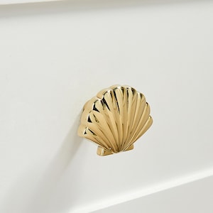Puede incluir: Un pomo de mueble de metal dorado con forma de concha marina y acabado pulido y reflectante. El pomo presenta crestas detalladas y elevadas que irradian desde la base hasta el borde exterior, montado sobre una superficie blanca lisa.