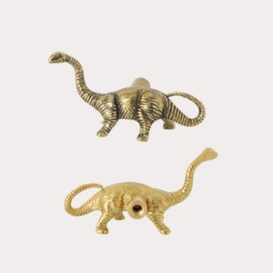 May include: Two brass-toned dinosaur-shaped cabinet knobs. Each knob features a long-necked sauropod design with a textured metallic finish and a cylindrical mounting post extending from the center of the dinosaur's back for attachment to furniture.