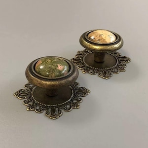 Puede incluir: Dos pomos redondos para armarios con bases de metal en tono latón antiguo que presentan placas traseras ornamentadas de estilo filigrana. Un pomo tiene una incrustación de piedra lisa moteada en verde y rosa, el otro un centro de piedra beige y marrón.