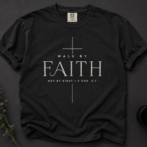 Puede incluir: Camiseta negra con las palabras "WALK BY FAITH" y "FAITH" en blanco, con una cruz. El texto "NOT BY SIGHT • 2 COR. 5:7" está debajo. La camiseta tiene mangas enrolladas y una etiqueta Comfort Colors.