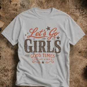 Op de afbeelding: Een lichtgrijs t-shirt met de tekst "Let's Go GIRLS GOOD TIMES GREAT VIBES" in vintage stijl. De tekst is in bruin- en oranje tinten met steraccenten. Het shirt heeft een ronde hals en korte mouwen.