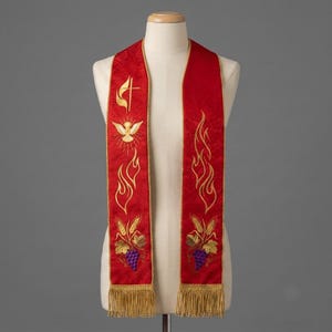 May include: A red liturgical stole featuring gold embroidered designs of a cross, a dove, flames, wheat stalks, and purple grapes. The stole is finished with gold fringe at the bottom edges and has a subtle tonal pattern throughout the fabric.