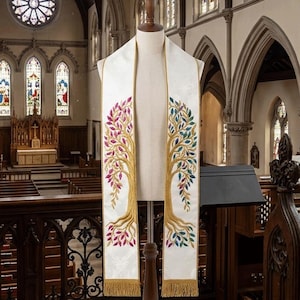 May include: A white liturgical stole featuring two embroidered trees with gold trunks and roots. The left tree has pink and purple leaves, while the right tree has blue and green leaves. The stole is finished with gold fringe at the bottom edges.