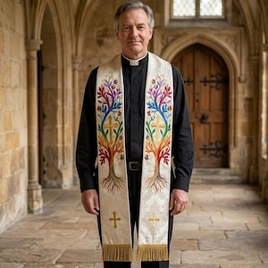 May include: A black clerical shirt worn under a cream-colored liturgical stole featuring embroidered rainbow-colored trees, gold crosses, and gold fringe at the bottom. The stole displays a symmetrical design with vibrant leaf patterns and roots.