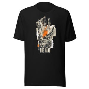 Può includere: Una t-shirt nera a maniche corte con una stampa grafica di una mano sovrapposta a ritagli di giornale e un globo. Il design include una macchia di vernice arancione brillante sul palmo e il testo ONE HAND stampato in bianco in basso.