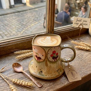 Puede incluir: Una taza de cerámica de forma cuadrada con esmalte reactivo, que presenta dos paneles ovalados con ilustraciones florales y un lazo. La taza contiene un café con leche con arte de espuma en forma de hoja. Una cuchara cobriza y espigas de trigo.
