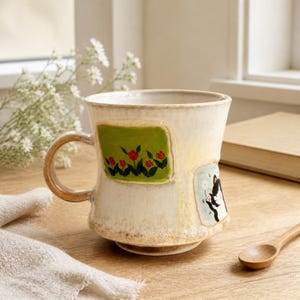 Könnte beinhalten: Ein Keramikbecher mit cremefarbener, texturierter Glasur, verziert mit einem handgemalten grünen Rechteck mit roten Tulpen und einem hellblauen Feld, das eine schwarz-weiße Katze zeigt. Der Becher hat einen breiten Rand und einen geschwungenen Henkel.