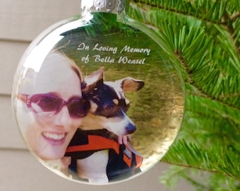 Op de afbeelding: Een transparante glazen kerstbal met een foto van een vrouw en een hond erin. De bal heeft een paars lint en de tekst "In loving memory of Bella Weasel" erop gedrukt.
