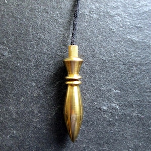 Op de afbeelding: Een messing slinger met een puntige bodem en een cilindrische bovenkant, opgehangen aan een zwart koord. De slinger heeft een glad, gepolijst oppervlak en een klassiek ontwerp. De achtergrond is een donker, getextureerd oppervlak.