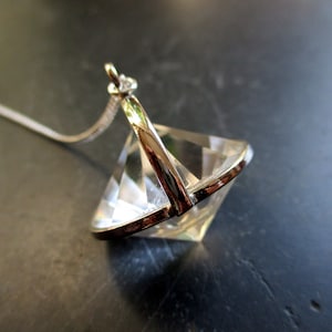 May include: A clear, faceted crystal pendant with a silver-tone metal frame and chain. The pendant is diamond-shaped, reflecting light. The chain is delicate and thin. The jewelry is displayed on a dark, textured surface.