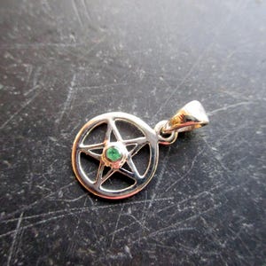 May include: A silver pendant with a pentagram design and a small green gemstone in the centre. The pendant has a loop for a chain. The jewellery is set against a dark, textured background.