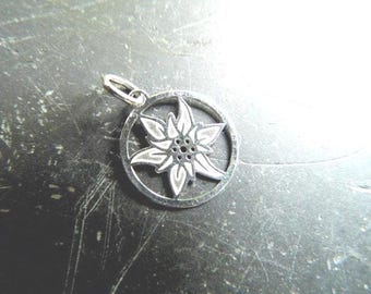 May include: A circular silver pendant featuring a cutout edelweiss flower design with six pointed petals and a central cluster of small dots. The charm includes a small silver jump ring attached to the top for hanging on a chain or cord.