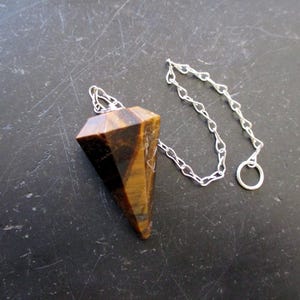 May include: A faceted tiger's eye pendulum with a silver chain and ring. The pendulum is a warm brown colour with darker brown stripes. The chain and ring are silver. The pendulum is used for divination and spiritual practices.