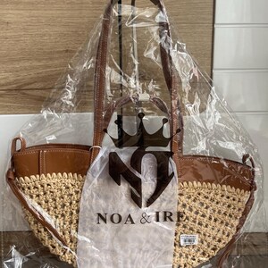 K&ouml;nnte beinhalten: Eine neue Tragetasche mit gewebtem Strohboden und braunen Lederakzenten. Die Tasche befindet sich in einer transparenten Plastikt&uuml;te mit dem Markennamen "NOA & IRE" aufgedruckt. Sie hat braune Ledergriffe.