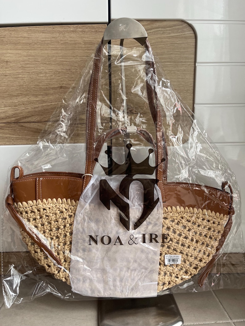 K&ouml;nnte beinhalten: Eine neue Tragetasche mit gewebtem Strohboden und braunen Lederakzenten. Die Tasche befindet sich in einer transparenten Plastikt&uuml;te mit dem Markennamen "NOA & IRE" aufgedruckt. Sie hat braune Ledergriffe.