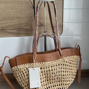 K&ouml;nnte beinhalten: Eine gewebte Stroh-Tragetasche mit braunen Lederbes&auml;tzen und -griffen. Die Tasche hat ein Etikett mit der Aufschrift "NOA & IRE". Die Tasche wird auf einem Metallst&auml;nder pr&auml;sentiert.