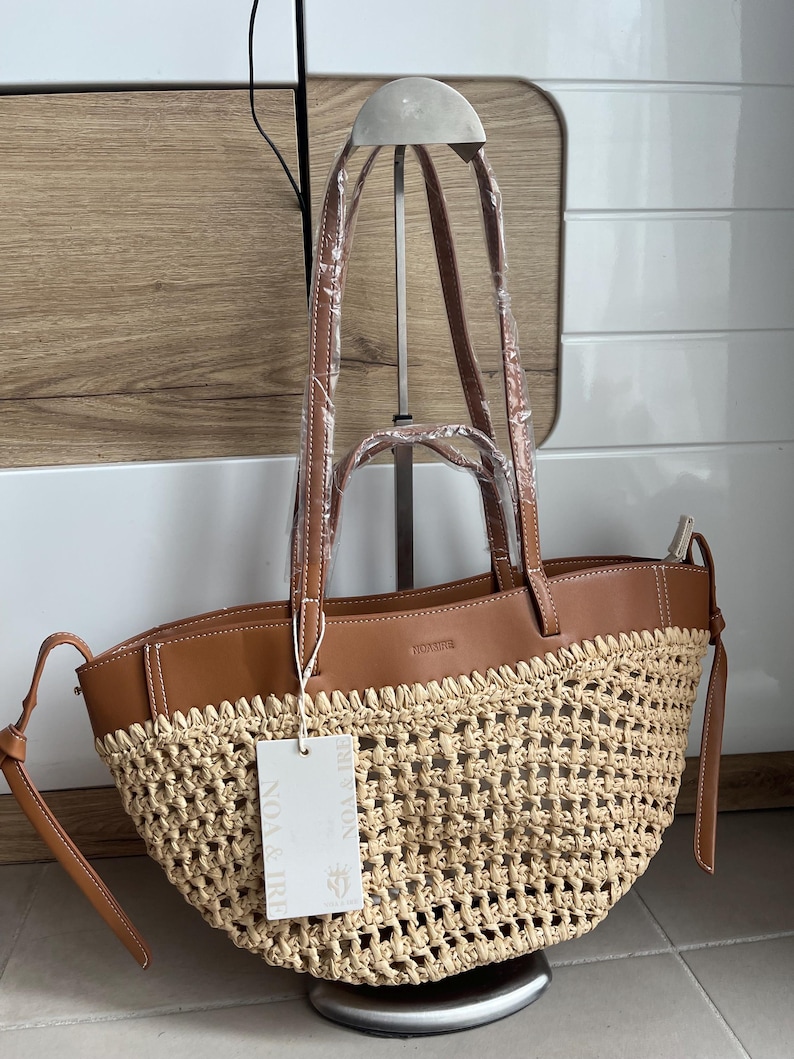 K&ouml;nnte beinhalten: Eine gewebte Stroh-Tragetasche mit braunen Lederbes&auml;tzen und -griffen. Die Tasche hat ein Etikett mit der Aufschrift "NOA & IRE". Die Tasche wird auf einem Metallst&auml;nder pr&auml;sentiert.