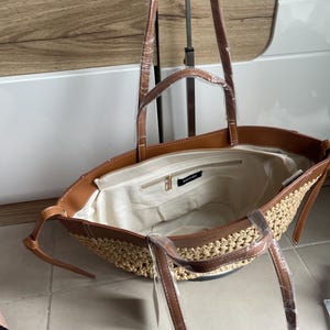 K&ouml;nnte beinhalten: Offene Tragetasche mit gewebtem Strohboden und braunen, leder&auml;hnlichen Verzierungen. Die Tasche hat lange Riemen und ein Innenfach mit Rei&szlig;verschluss. Das Innenfutter ist hellbeige. Der Markenname "NOAR & BEE" ist sichtbar.