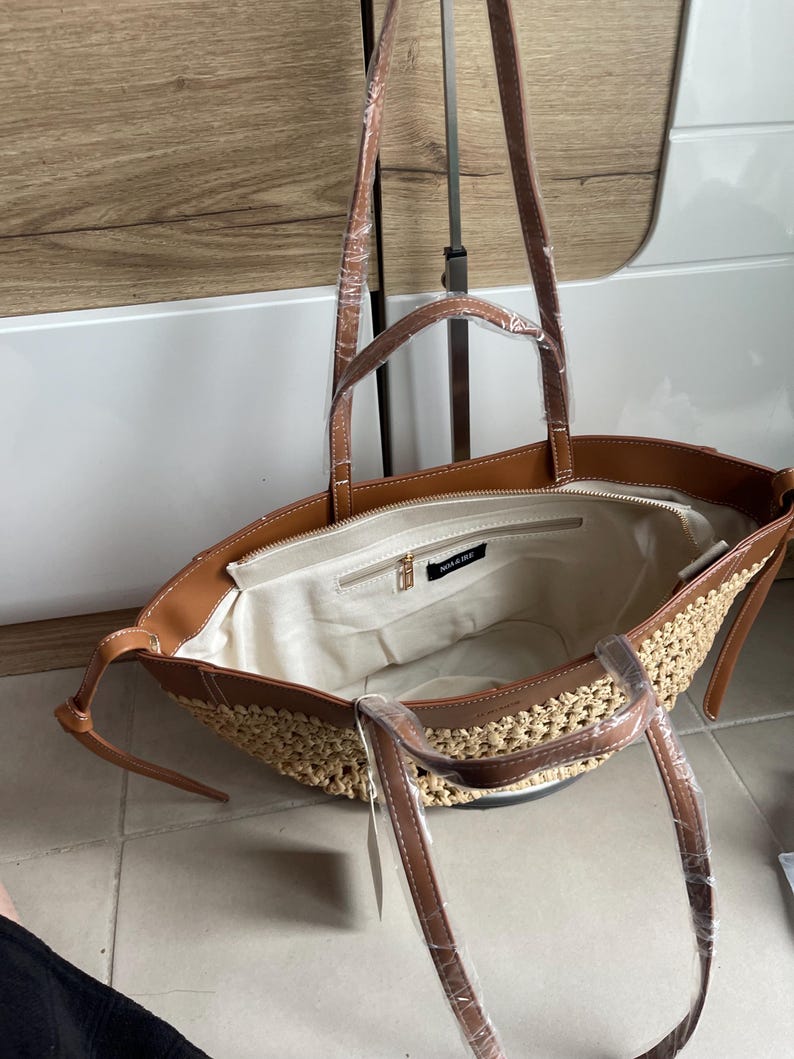 K&ouml;nnte beinhalten: Offene Tragetasche mit gewebtem Strohboden und braunen, leder&auml;hnlichen Verzierungen. Die Tasche hat lange Riemen und ein Innenfach mit Rei&szlig;verschluss. Das Innenfutter ist hellbeige. Der Markenname "NOAR & BEE" ist sichtbar.