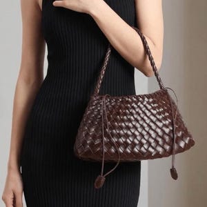 Peut inclure: Un sac à main marron foncé présentant un motif en cuir tressé sur tout le corps. Le sac comprend une bandoulière en cuir tressé et de fins cordons de serrage en cuir sur les côtés qui se terminent par de petites languettes en cuir arrondies.