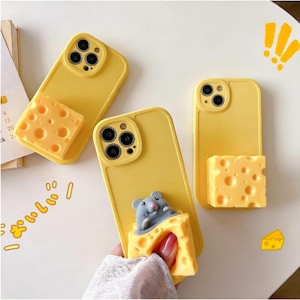 May include: Yellow phone cases with a cheese-shaped design. One case features a gray mouse peeking out of the cheese. The cases have a camera cutout and are on a white surface.