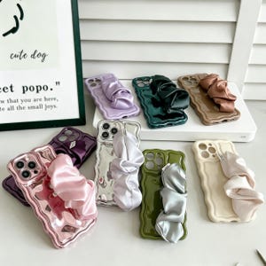 Könnte beinhalten: Eine Sammlung von Smartphone-Hüllen mit gewelltem Rand in verschiedenen Farben wie Lavendel, Dunkelgrün, Beige, Metallic-Rosa, Silber, Olivgrün und Creme. Jede Hülle verfügt über ein passendes Stoffband im Scrunchie-Stil auf der Rückseite.