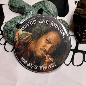 Puede incluir: Un botón redondo con una foto en blanco y negro de un hombre fumando un cigarrillo. El texto en el botón dice "knives are knives, meat's meat."