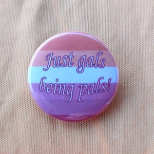 Pode incluir: Um botão redondo com um design listrado rosa, branco e laranja. O botão tem o texto "Just gals being pals!" em escrita cursiva branca.