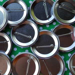 K&ouml;nnte beinhalten: Nahaufnahme der R&uuml;ckseiten von zehn silbernen Metall-Knopfn&auml;geln mit Sicherheitsnadeln. Die N&auml;gel sind in einem Halbkreis auf einem gr&uuml;n-wei&szlig; gepunkteten Hintergrund angeordnet.