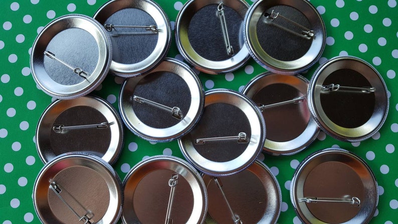 K&ouml;nnte beinhalten: Nahaufnahme der R&uuml;ckseiten von zehn silbernen Metall-Knopfn&auml;geln mit Sicherheitsnadeln. Die N&auml;gel sind in einem Halbkreis auf einem gr&uuml;n-wei&szlig; gepunkteten Hintergrund angeordnet.