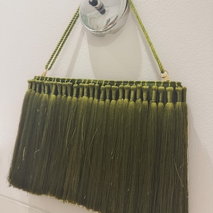 May include: A rectangular olive green clutch bag featuring a dense layer of long, matching green fringe covering the entire front surface. The bag includes a twisted green rope handle attached to gold-toned hardware, allowing it to hang vertically.