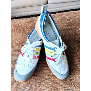 May include: A pair of white leather golf shoes with blue, yellow, green, and red accent stripes along the lace-up area. The shoes feature a grey toe cap, white laces, silver-tone eyelets, and the text Sesto Golf by Sherry printed on the insole.