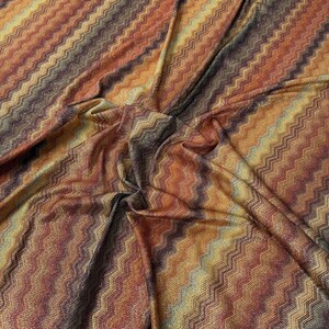 Puede incluir: Una tela ligera con un patrón de chevrón repetitivo en tonos cálidos de naranja quemado, burdeos profundo, amarillo dorado y marrón oscuro. El material tiene una textura fina similar al tejido de punto con hilos metálicos sutiles.