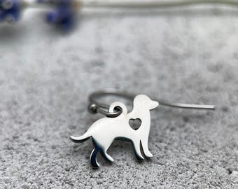 Puede incluir: Un pendiente de metal plateado con la silueta recortada de un perro que presenta un pequeño orificio en forma de corazón en la zona del pecho. El dije está unido a un gancho de metal curvado para el lóbulo de la oreja.