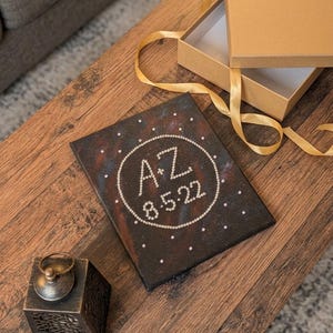 May include: A rectangular canvas with a dark, galaxy-inspired background featuring a circular border of small white beads. Inside the circle, the letters A+Z and the date 8.5.22 are spelled out in white beads. The canvas is accompanied by a gift box.